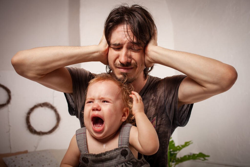 Papa qui ne supporte pas les pleurs de bébé : c'est normal, voici pourquoi et comment tenir 2 Homme se bouchant les oreilles ou montrant des signes de détresse face aux cris d'un bébé.